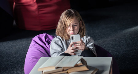 A little girl lies on a pouf and uses a smartphone instead of reading a book.の写真素材
