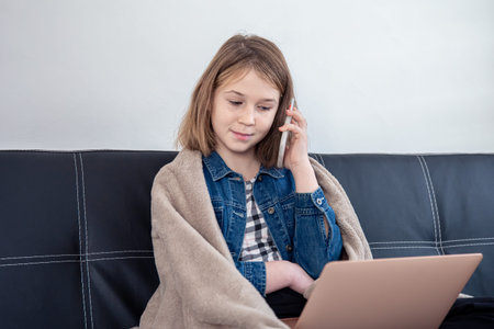 A teenage girl is sitting on the couch at a laptop and talking on the phone.の写真素材