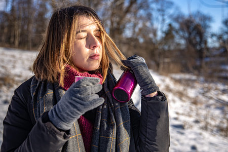 A young woman enjoys a hot drink from a flask on a walk in winter.