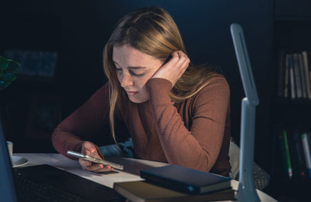 A young woman with a smartphone in her hands works late at night.