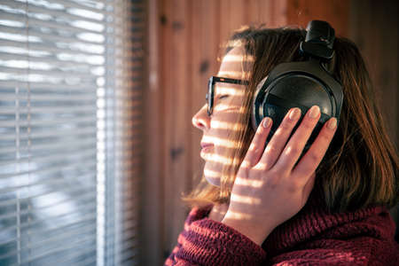 A woman with headphones looks through the blinds at the early morning sunlight.の写真素材