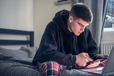 A teenager guy sits in a room on a bed and uses a laptop.の写真素材