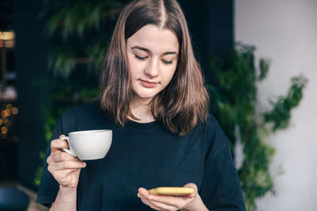 Teenage girl with a cup of tea and a smartphone in her hands.の写真素材