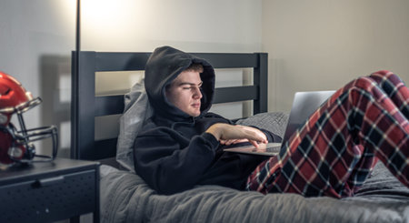 A teenager guy sits in a room on a bed and uses a laptop.