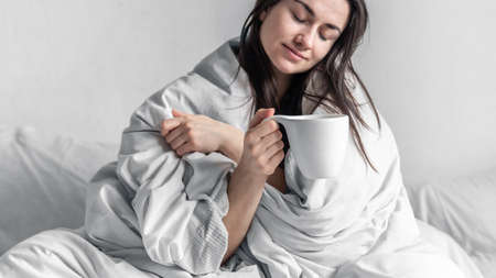 A beautiful young woman in a white bed with a cup of coffee.の写真素材