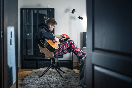 A young man plays the acoustic guitar in his room at home.