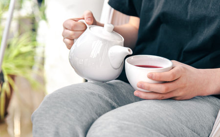A white cup of tea and a teapot in the hands of a woman sitting in a hammock.の写真素材
