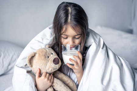 A little cute girl in bed with a plush and a glass of milk.の写真素材