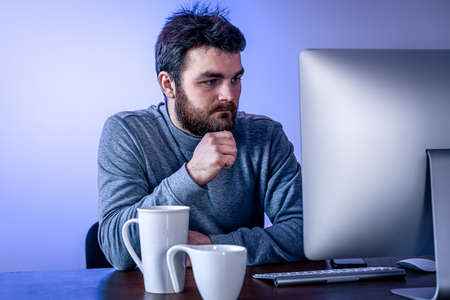 Tired man sits in front of a computer with a cup of coffee, colored lighting.の写真素材