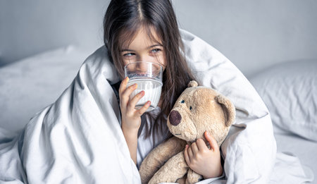 A little cute girl in bed with a plush and a glass of milk.の写真素材