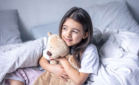 Sleepy little girl with her favorite teddy bear in bed in the morning.の写真素材