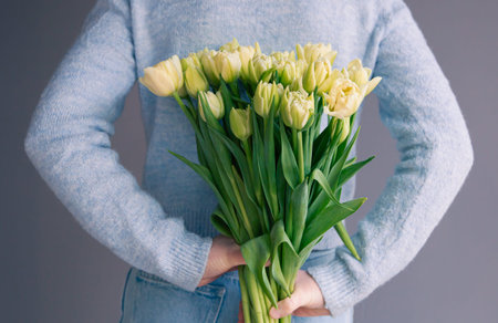 A woman holds a bouquet of tulips behind her back on a gray background.