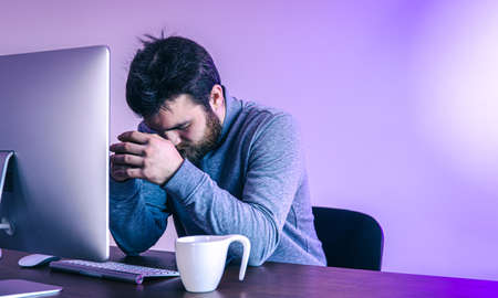 Tired man sits in front of a computer with a cup of coffee, colored lighting.の写真素材