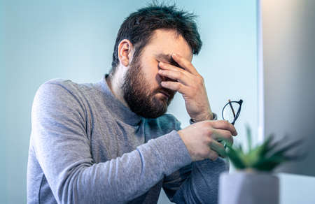 A tired man rubs his eyes in front of a computer screen.の写真素材