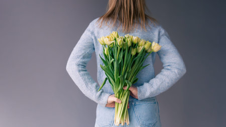 A woman holds a bouquet of tulips behind her back on a gray background.