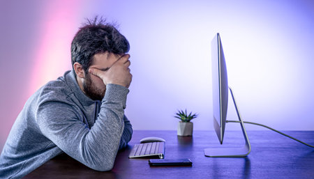 A tired man sits in front of a computer, covering his face with his hands.