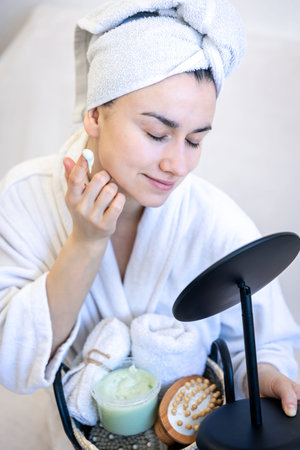 A young woman applies cream on her face, looks in the mirror.の写真素材