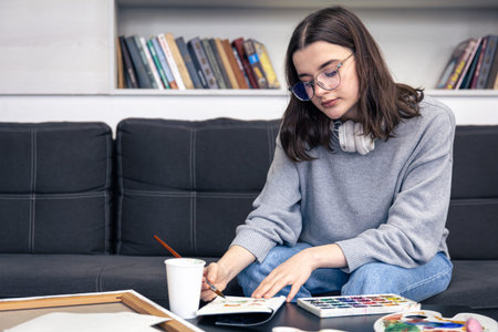 A stylish young woman draws with paints in the home interior.