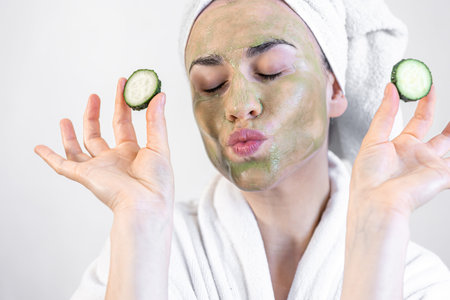Young woman with a green face mask and fresh cucumbers in a white bathrobe.の写真素材