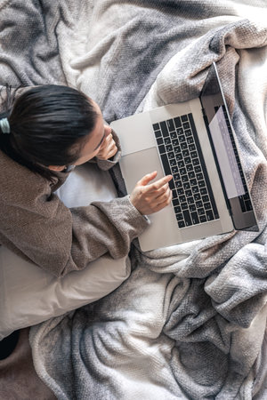 A woman working on a laptop while lying in bed, top view.