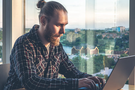 A man with a beard works at a computer, sitting in the office by the window.