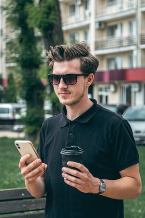 A young man in sunglasses with smartphone and coffee in the city.