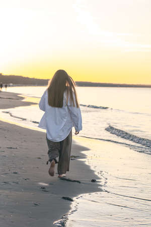 A woman walks along the sea beach at sunset, view from the back.の写真素材