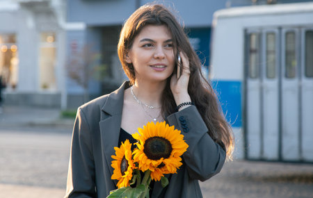 A young woman with a bouquet of sunflowers is talking on the phone at sunset.