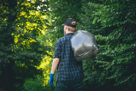 A young man in gloves and with a garbage bag cleans up in the forest.