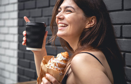 A young woman with a croissant and a cup of coffee against a black brick wall.