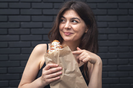 A young woman with a croissant and a cup of coffee against a black brick wall.