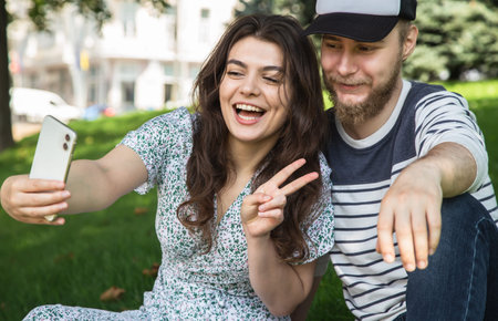 A guy and a girl take a selfie sitting on the grass in the park.