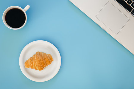 Croissant, cup of coffee and laptop on a blue background, flat lay.の写真素材
