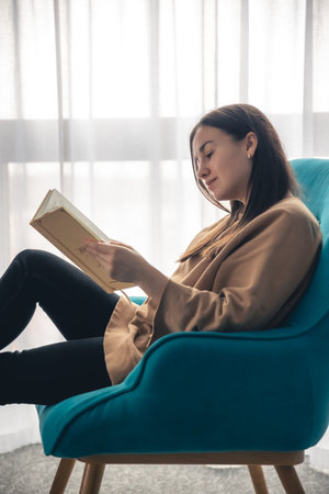 A young woman is reading a book while sitting on an armchair by the window.