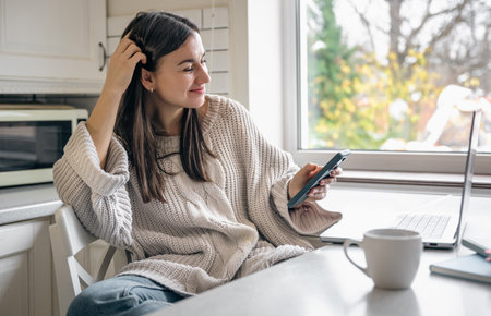 A young woman is sitting in the kitchen with a smartphone and a laptop.の写真素材