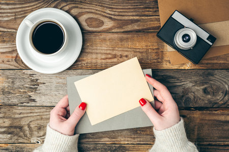 An envelope and a blank sheet of paper in female hands on a wooden background with a retro camera and a cup of coffee, top view.の写真素材
