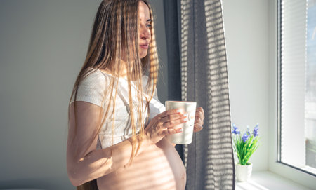 An attractive pregnant woman with a cup of tea at the window in the morning, the concept of women's health and the expectation of a child.の写真素材