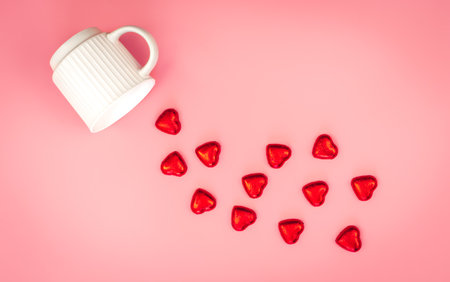 A cup and scattered heart-shaped chocolate candies in red foil on a pink background, flat lay.の写真素材
