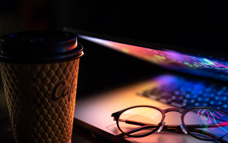 A laptop half closed in the dark with colourful glow, cup of coffee and glasses.の写真素材