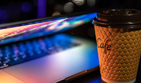 A laptop half closed in the dark with colourful glow and cup of coffee.の写真素材