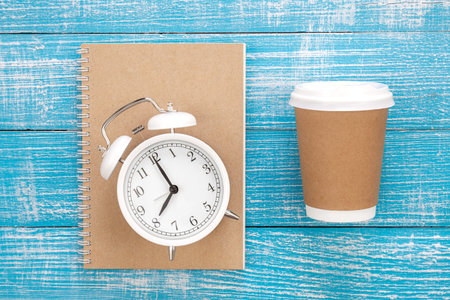Paper disposable glass, notepad and alarm clock on a wooden background.