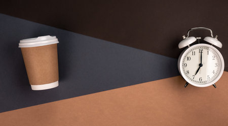Disposable paper cup and white alarm clock on paper background, top view.