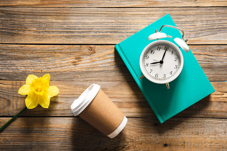 Book, alarm clock, paper cup and flower on wooden background, top view.