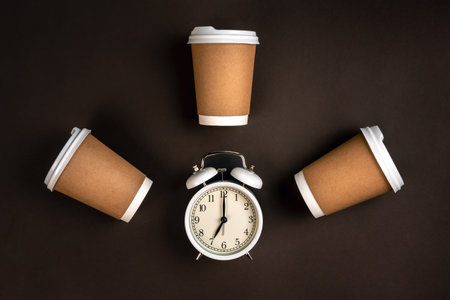 White alarm clock and craft disposable cups on a dark background, top view.の写真素材