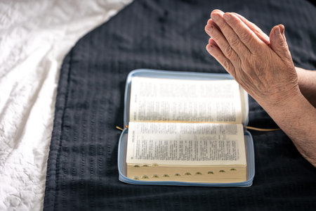 The hands of an elderly woman folded in prayer in front of a book of the Bible.