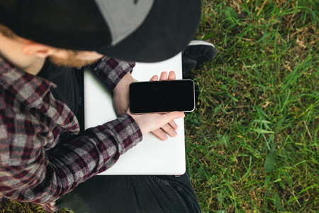 A man is sitting on the grass with a laptop and a smartphone in his hands.の写真素材