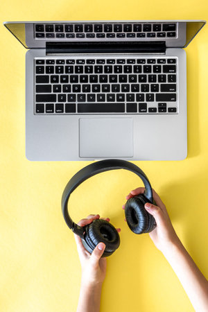Headphones in the hands of a child and a laptop on a yellow background.