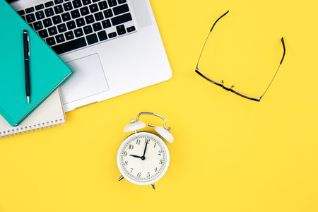 Laptop, alarm clock and glasses on a yellow background.
