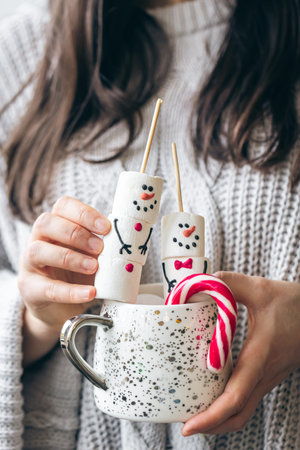 Cup of hot drink with marshmallow snowmen and candy in female hands.