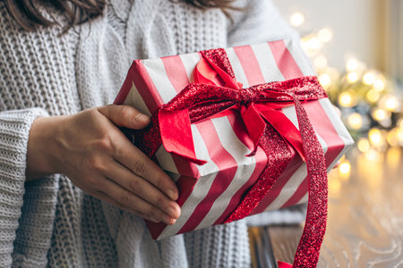 A holiday gift with a red bow in female hands on a blurred one with garlands.の写真素材